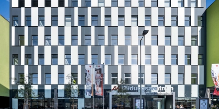 Frontansicht des Eingangbereichs inklusive Vorplatz. Die Fassade mit Glas- und Metallelementen rhythmisierend abwechselnd in Schwarz-Weiß.