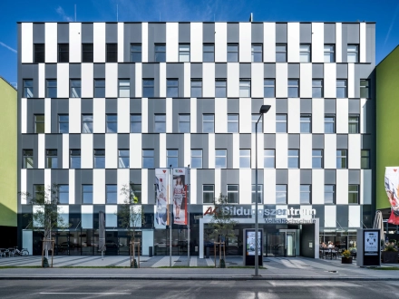 Frontansicht des Eingangbereichs inklusive Vorplatz. Die Fassade mit Glas- und Metallelementen rhythmisierend abwechselnd in Schwarz-Weiß.